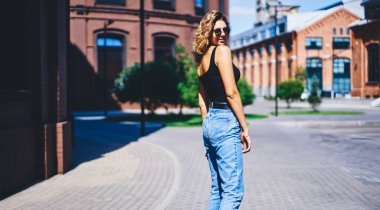 Güneşli bir günde güneş gözlüğü ve güneş gözlüğü takan, gülümseyen ve arkasını kollayan, bronz tenli, iyimser genç bir kadının yan görüntüsü.