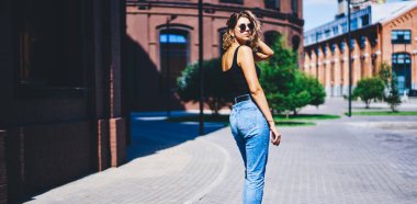 Gündelik giysiler ve güneş gözlükleriyle saçlarına dokunan ve kaldırım boyunca yürürken omzunun üstünden bakan çekici genç kadın modelin arka görüntüsü.