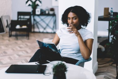 Sakin, genç, etnik bir kadın, günlük kıyafetlerini giymiş dergi okuyor ve rahat bir sandalyede oturmuş, gün ışığında kameraya bakarken bir fincan kahvenin tadını çıkarıyor.
