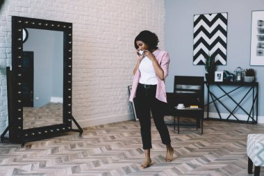 Çağdaş stüdyoda aynanın yanında dikilirken cep telefonuyla konuşan gündelik kıyafetli, çıplak ayaklı, Afro-Amerikan genç bir kadın.