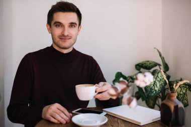 Olumlu genç adam ahşap masada oturmuş elinde bir fincan sıcak kahveyle akıllı telefon ve defterle çalışırken ve kameraya bakarken.