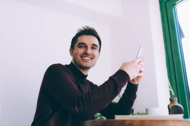 Hafif bir kafedeki ahşap masada oturmuş kameraya bakarken sıradan süveter giyen neşeli genç adamın yan görüntüsü.