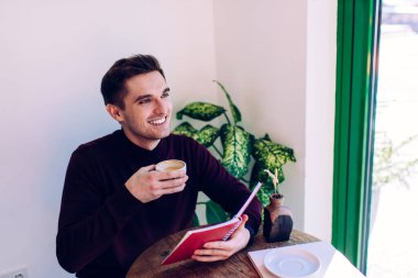 Öğrenim ve öğrenim için eğitim kitabı tutan neşeli Kafkasyalı öğrenci, kolej ödevlerini yaparken gülümseyen, planlayıcı not defteri ve kafein içeceğiyle neşeli bir erkek.