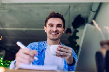 Düşük açılı gülümseyen genç bir erkek bir fincan sıcak içecekle kaldırımdaki kafede dizüstü bilgisayar kullanarak not alıyor.