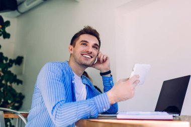 Ellerinde dijital tabletle neşeli bir binyıl blogcusu sahte dizüstü bilgisayarlı masaya gülüyor, modern teknolojiyi sosyal ağ ve web taraması için kullanıyor.