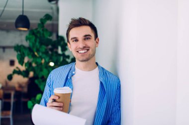 Gündelik giyinen, gülümseyen, beyaz duvarın yanında duran ve modern ofiste bir fincan kahve ve belgelerle mutlu bir erkek.