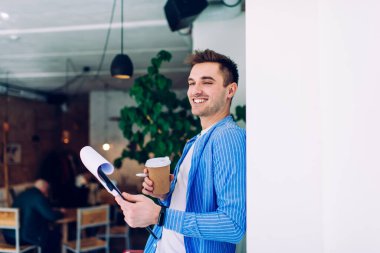 Kahve dükkanında öğrenilmek için gülümseyen tek kullanımlık kahve fincanı ve kağıt klasörlü neşeli erkek, mavi gömlekli, kafein içeceği tutan mutlu beyaz adam, duvarın içinde gülüyor.