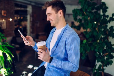 Kafede dikilip Tim Ein 'i boş vakitlerinde geçirirken rahat giyinen, gülümseyen ve akıllı telefonlara bakan genç adamın yan görüntüsü.