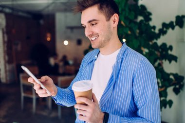 Mavi gömlekli mutlu beyaz adam cep telefonuyla kahveyle ilgili çevrimiçi geri bildirim yazıyor, paket servis bardağıyla sosyal ağlarda hücresel iletişimin keyfini çıkaran neşeli erkek blogcu