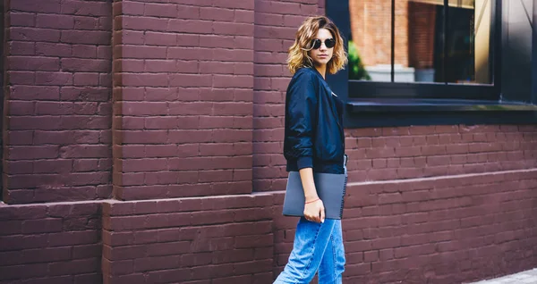 Gündelik kıyafetleri ve güneş gözlükleriyle modern binanın yanında elinde not defteriyle dikilip kameraya bakan modaya uygun genç bir kadın.