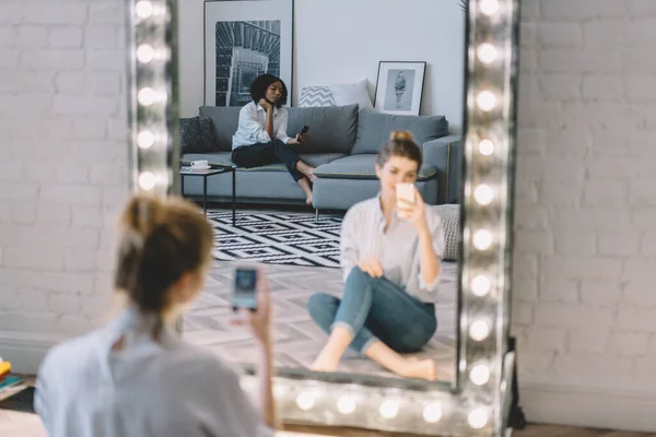 Woman sitting at illuminated mirror and taking selfie while African American girlfriend resting on sofa and browsing in cozy loft style living room