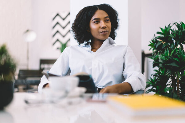 Positive young ethnic female in casual clothes with book sitting at table with cup of hot drink and notebooks at modern apartment and looking away