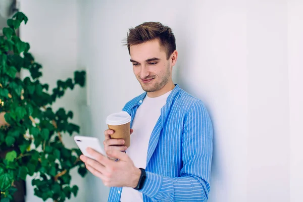 Mavi gömlekli mutlu beyaz adam, cep telefonuyla kahveyle ilgili çevrimiçi geri bildirim yazıyor, paket servis bardağıyla sosyal ağlarda hücresel iletişimin keyfini çıkaran yakışıklı erkek blogcu.