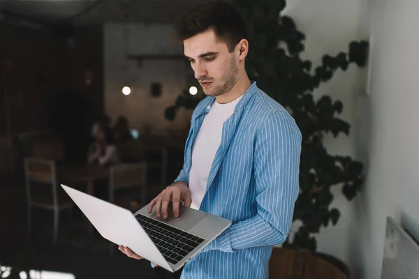 Serious male freelancer in casual wear standing with laptop and using touchpad while working on project in cafe on blurred background