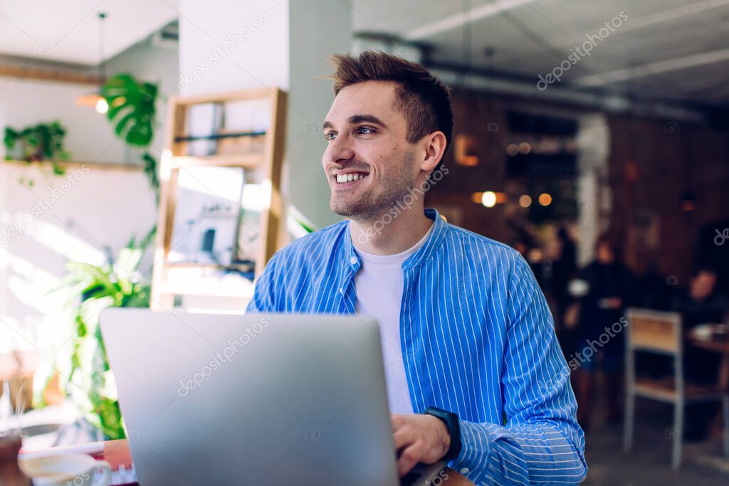 Funny male programmer dressed in smart casual blue shirt smiling during remote work on laptop computer, cheerful Caucasian software developer 20 years old with digital netbook laughing indoors