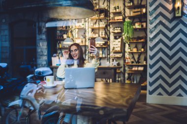 Pencere manzarası, sarı kazaklı ve gözlüklü güzel esmer kadının laptopuyla kafede otururken selfie çekmesi.