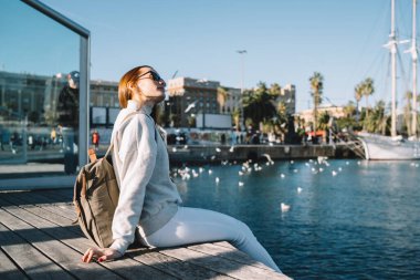 Moda kıyafetli bir kadın turist liman kenarında oturmuş, seyahat masraflarını düşünüyor, güneş gözlüklü güzel beyaz kadın şehir rıhtımında eğleniyor.