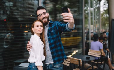 Gündüz vakti modern kafenin yanında durup cep telefonuyla selfie çeken mutlu bir adam ve kadın.