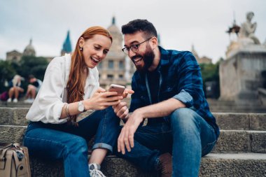 Genç ve mutlu bir çift, tarihi binanın önünde oturmuş gülerken akıllı telefondan eğlenceli görüntüler izliyorlar.