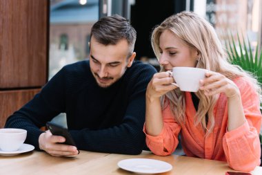 Neşeli adam kız arkadaşına akıllı telefon ekranı gösteriyor. Bu arada kadın akıllı telefona bakıyor ve kaldırımdaki kafede çay içiyor.