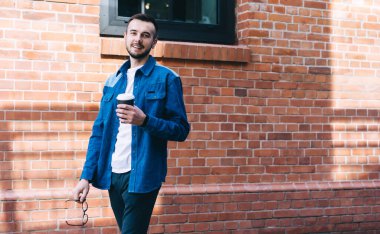 Kotlu mutlu genç adam kameraya gülümserken elinde kahve bardağıyla şehirde tuğla duvar boyunca yürüyor.