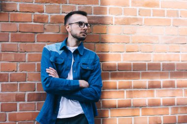 Ciddi sakallı, gündelik kıyafetleri ve gözlükleriyle tuğla duvarın yanında dikilen ve güneşli bir günde düşünceli bakışları kaçıran genç bir adam.