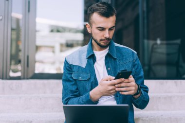 Kot ceketli ve beyaz tişörtlü dalgın genç bir erkek modern binanın girişinde cep telefonundan bilgi arıyor.