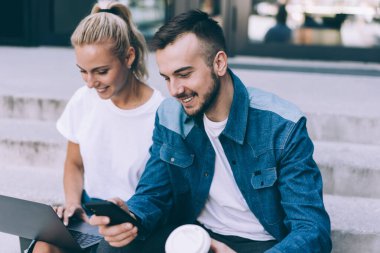 Genç gülümseyen sakallı bir adam, bir fincan kahve içip cep telefonlarını karıştırıyor ve merdivenlerde oturmuş neşe içinde ağlarda sörf yapıyor.