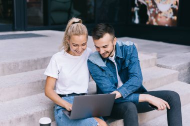 Basamaklarda kağıt kahve içip gülümserken dizüstü bilgisayarda gezinen genç kadın ve erkek pozitifler.