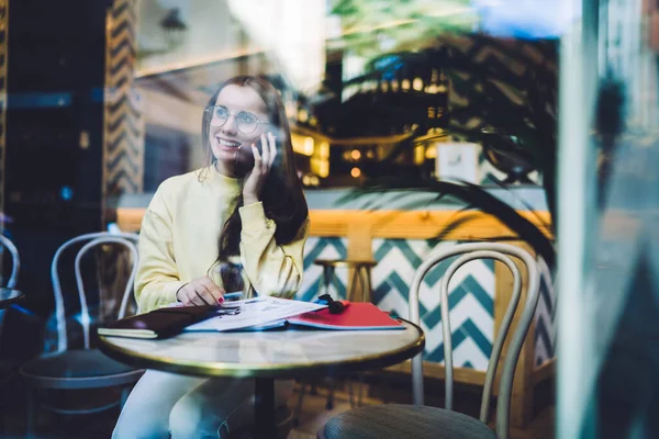 Moda gözlüklü, parlak sarı tişörtlü, telefonda gülümseyen, rahat, modern bir kafede oturan alımlı genç bir kadının yan görüntüsü.