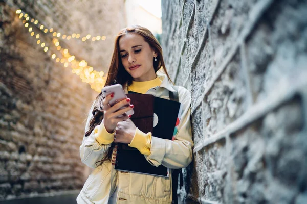 バルセロナ市内の通りに立っている間 スマートフォンや古い壁にもたれてノートを使用して美しい若い女性の低角度 — ストック写真