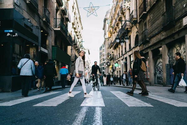 Side view of pretty young female with notebooks walking on crosswalk with crowd and looking away on narrow old street in Barcelona City