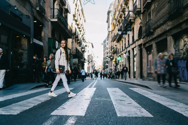 Side view of positive young woman with notepads crossing asphalt road on old narrow street during walk in Barcelona City