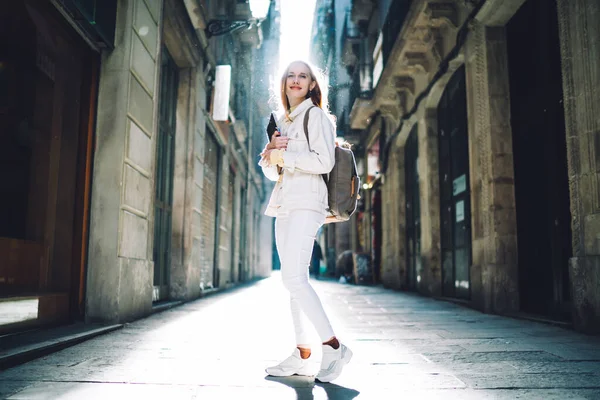 Inspired smart woman in white clothes with books and backpack walking around street between ancient buildings and houses in Barcelona