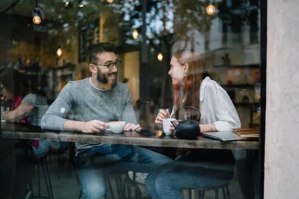 积极的年轻夫妇在白天休息时坐在舒适的咖啡店里 一边喝着热饮 一边互相微笑 — 图库照片