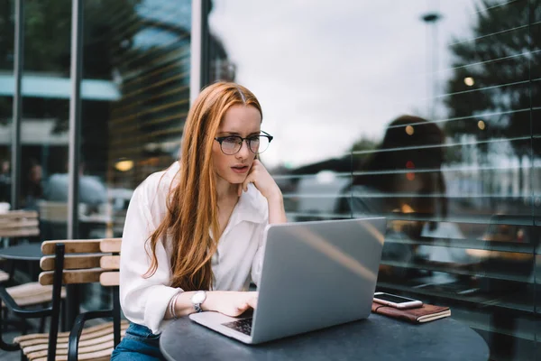 认真的女性 戴着眼镜 坐在现代咖啡馆外的桌子旁 一边用笔记本电脑键盘打字 一边做远程项目 — 图库照片