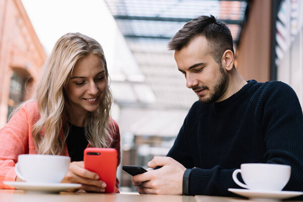 Улыбающиеся кавказские коллеги-женщины и мужчины делятся медиа-контентом и файлами на смартфоне через 4G связь отдыхая с педиками в кафе, веселые молодая женщина и мужчина ищут информацию в Интернете