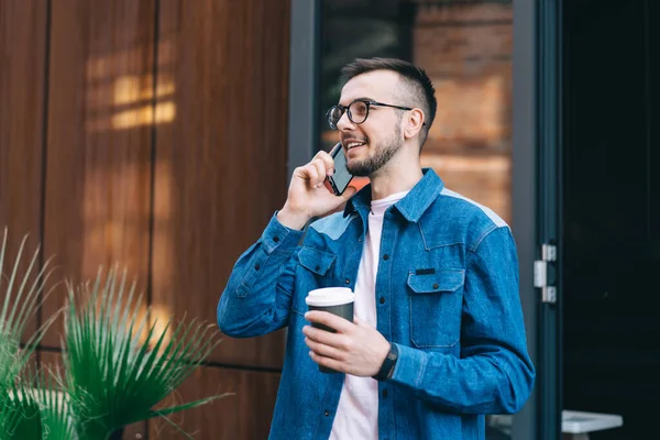 Smiling male hipster in casual clothes and glasses standing with cup of coffee and communicating on mobile phone in street