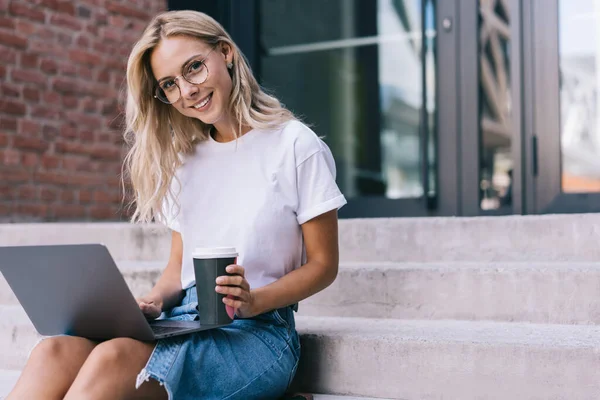 평상복 차림으로 금발의 여성을 낮에는 계단에 앉아서 컴퓨터로 일하면서 커피를 — 스톡 사진