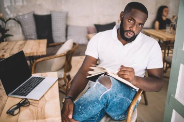 Ciddi koyu tenli erkek blog yazarının portresi yayıncılık için makale yazmaya odaklanmış, yakışıklı 20 'li yaşlarda Afro-Amerikan gazeteci ofisteki dizüstü bilgisayarın yanında oturan notlar alıyor.