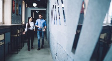 Beyaz bluzlu, gülümseyen bir kadın, modern iş yerinde günlük kot pantolonlu ve dizüstü bilgisayarı kapalı sakallı bir erkekle konuşuyor.