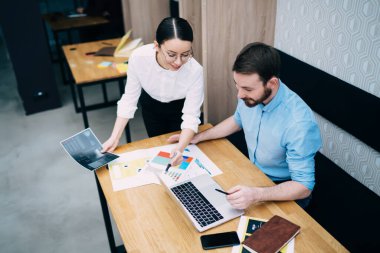 Resmi olarak memnun olmuş genç bayan muhasebeci iş arkadaşına belgeler ve elinde kalemle dizüstü bilgisayarla yardımcı oluyor ve çalışma alanına gülümsüyor.