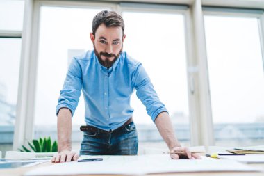 Aşağıdaki yakışıklı, yetişkin, sakallı, kaşlarını kaldıran ve gülümseyen bir çalışandan iş yerinde pencere duvarına yaslanıp, modern açık uzay ofisindeki kameraya bakarken.