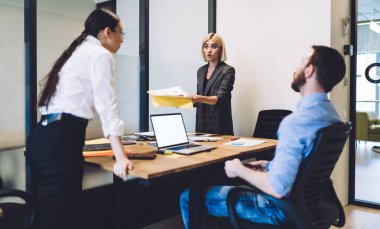 Sakin adam dizüstü bilgisayarla masada oturuyor ve iki kendine güvenen iş kadınının ofis odasında durup evrak işlerini konuşmalarını izliyor.