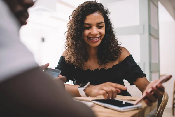 Cheerful female with curly hair showing smartphone device with browsed streams website enjoying live communication with male friend, cheerful girl networking internet via cellphone technology