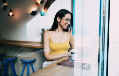 Optik gözlüklü neşeli yeniyetme kız göz düzeltmesi için çalışma alanında çalışıyor ve gülüyor, klasik gözlüklü mutlu beyaz kız öğrenci öğrenme için zaman harcıyor.