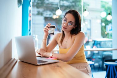 Modern dizüstü bilgisayar uygulaması üzerinden online webinar videolar izlemek için kahveyle çekici kadın kullanıcı, sosyal medya ağlarında klasik göz giysisi yayınında milenyum hipster kızı