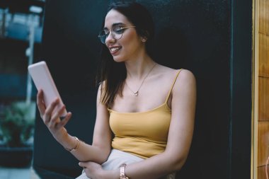 Mutlu beyaz kadın cep telefonundan mesaj gönderiyor ve bilgi okuyor. Açık havada dinleniyor. Klasik göz giysisiyle gülümseyen kadın akıllı telefon teknolojisinde pozitif video izliyor.
