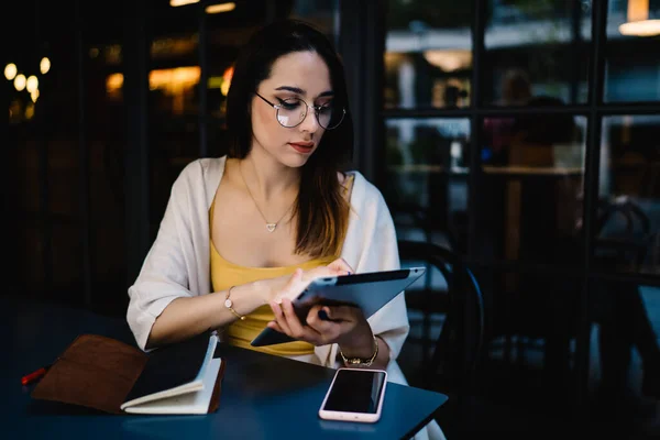 Millennial woman in spectacles sending sms messages via modern touch pad using 4g wireless in street cafe, female blogger checking online web statistics of shared influence video publication