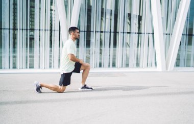 Çömelme hareketleri yapan kararlı erkek sporcu sabah antrenmanı sırasında fiziksel dinlenmenin keyfini çıkarıyor, kaslı sporcu enerji elde etmek için ısınma egzersizleri yapıyor.
